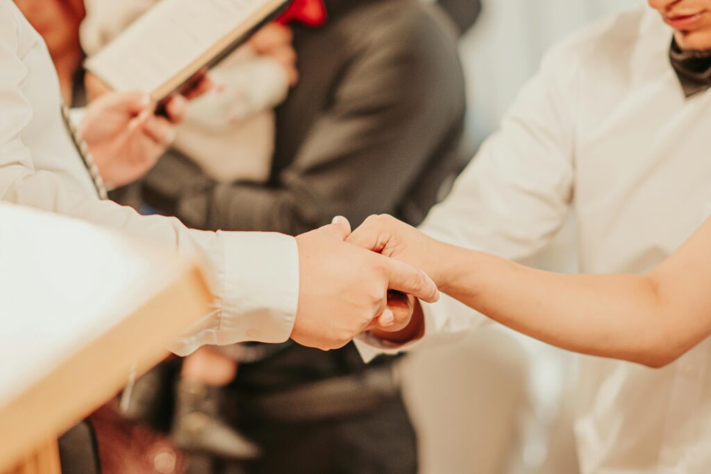 Unrecognizable priest with bible holding hands of anonymous bride and groom while taking vows during wedding ceremony with guests on blurred background