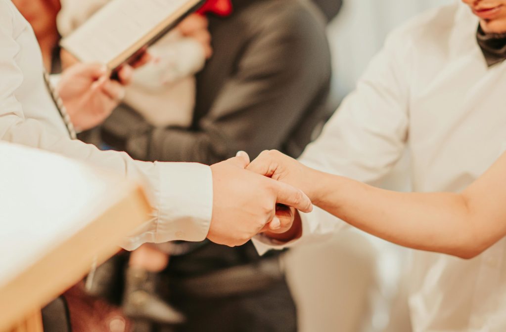 Unrecognizable priest with bible holding hands of anonymous bride and groom while taking vows during wedding ceremony with guests on blurred background