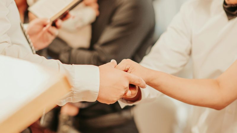 Unrecognizable priest with bible holding hands of anonymous bride and groom while taking vows during wedding ceremony with guests on blurred background